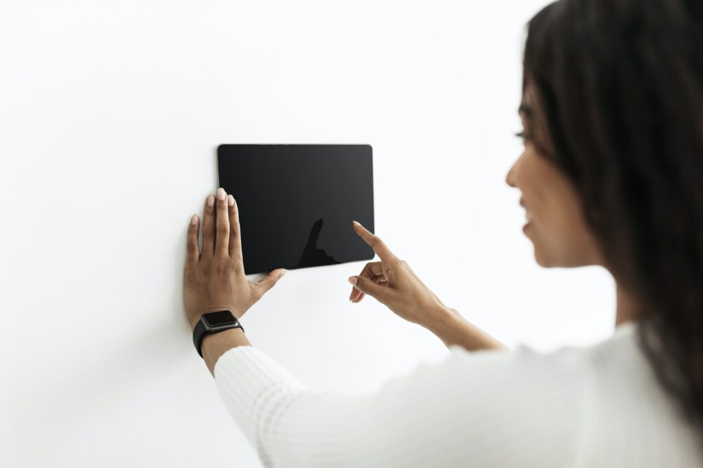 Young black lady controlling house with digital touch screen panel installed on white wall in the