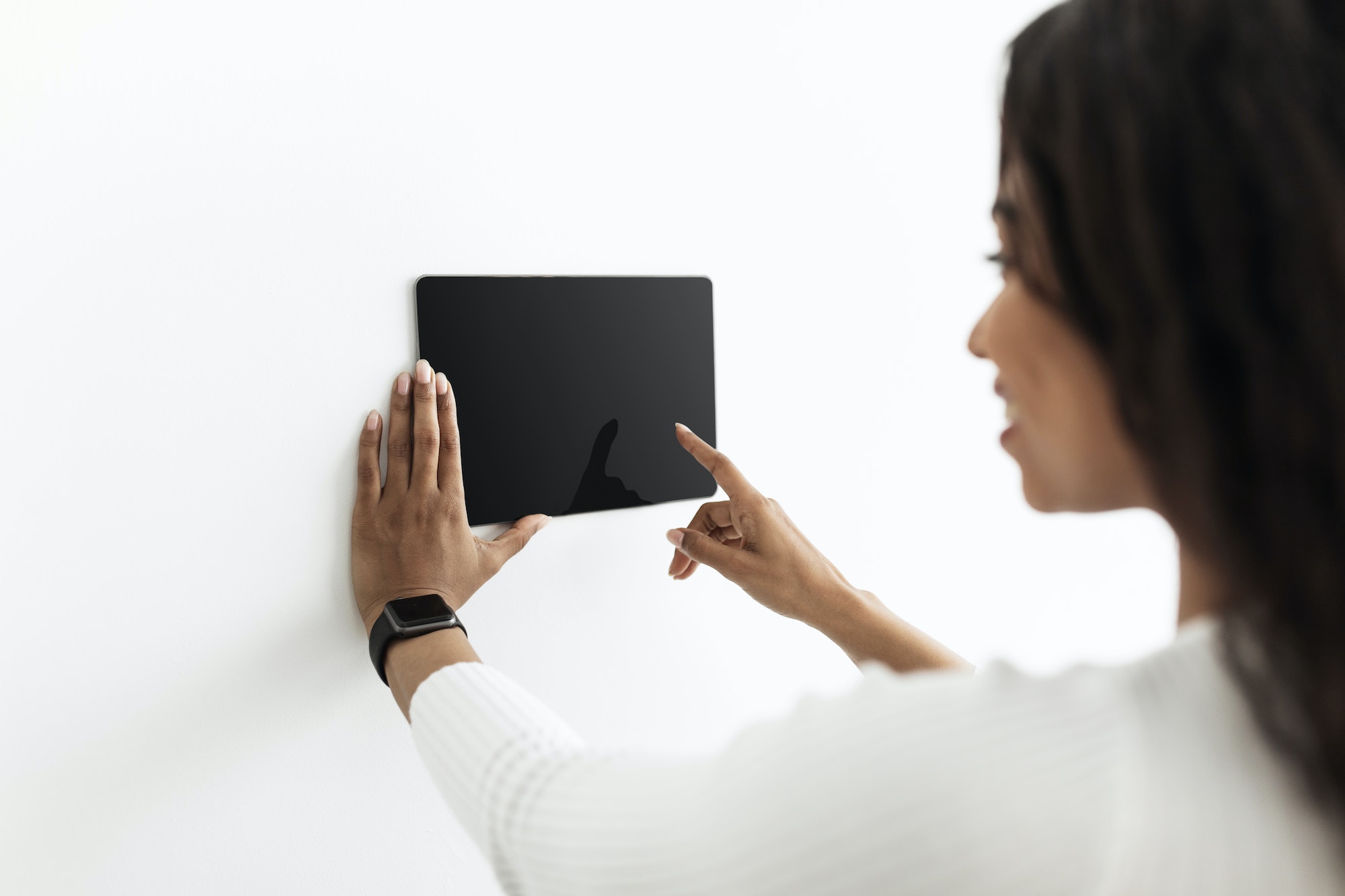 Young black lady controlling house with digital touch screen panel installed on white wall in the